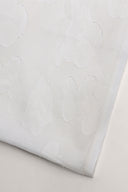 Gros plan sur l'angle plié du tissu écru, laissant apparaître le motif nuage en relief tissé et la lisière effilochée.