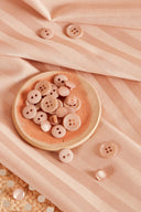 Tissu satin rayé rose poudré plié, avec une coupelle en céramique terracotta contenant des boutons roses nacrés.
