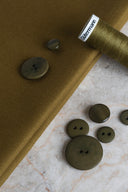 Tissu gabardine léger vert olive doré, présenté avec une bobine de fil Gütermann et des boutons assortis sur un plan de travail en marbre.