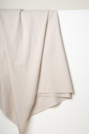 Tissu gabardine légère beige argile drapé en plis superposés en pointe, lisière effilochée visible.