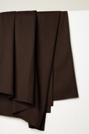 Chocolate brown light gabardine fabric draped in overlapping folds to a point, frayed selvedge visible.