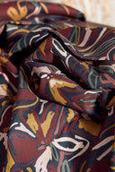 Tissu Hilma bordeaux plié, avec un dessous-de-plat en osier tressé contenant des boutons moutarde, bordeaux, sauge et roses.
